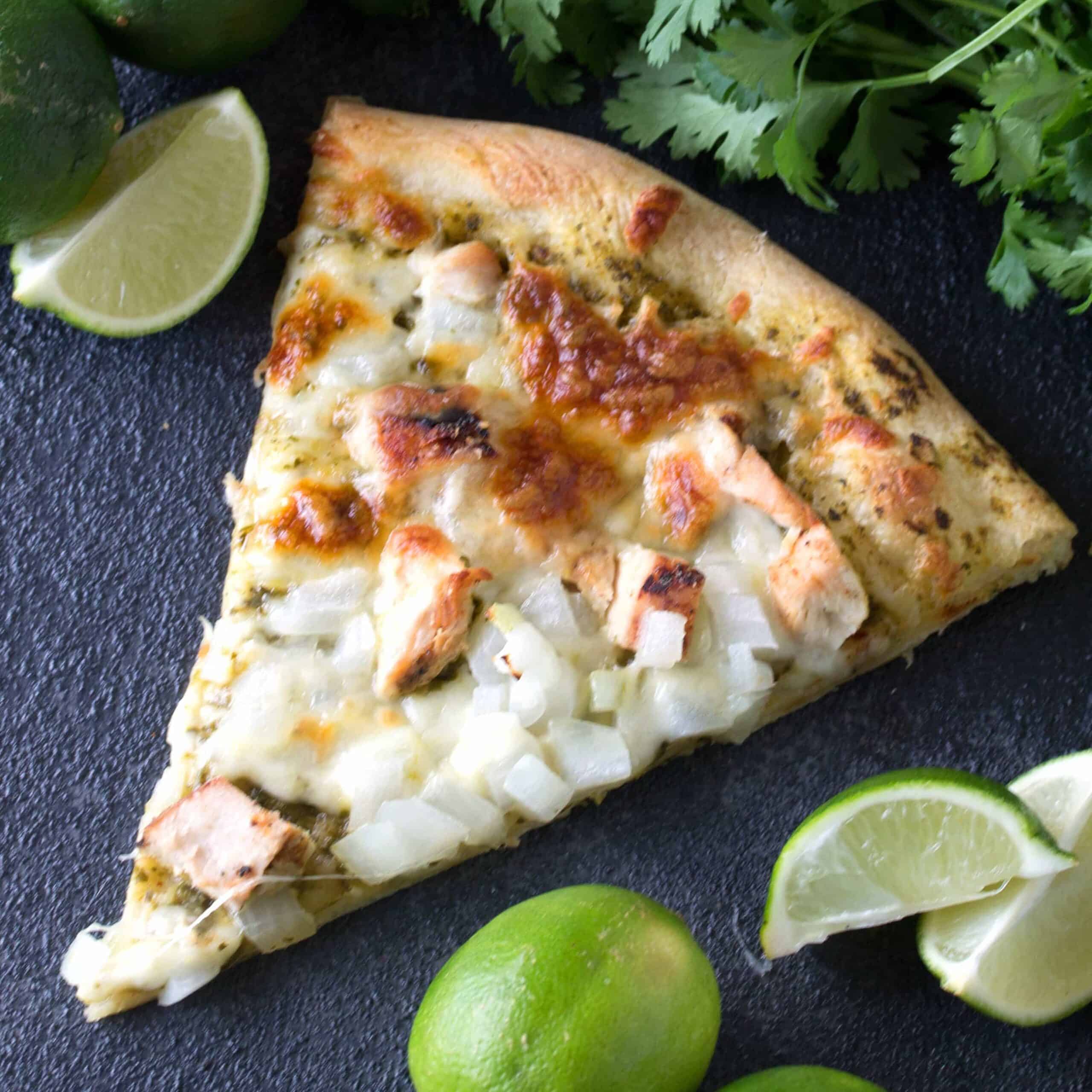 Slice of Cilantro Lime Chicken Pizza on a black countertop surrounded by lime wedges.