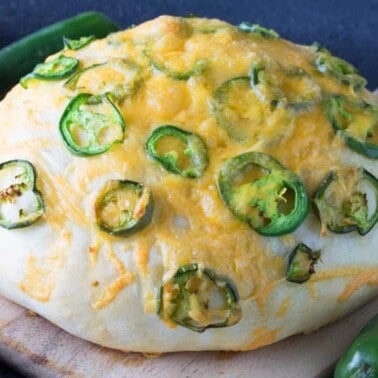Angled view of a loaf of artisan Jalapeno Cheese Bread on a wood cutting board surrounded by jalapeño peppers.