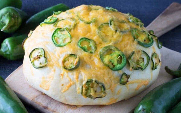 Angled view of a loaf of artisan Jalapeno Cheese Bread on a wood cutting board surrounded by jalapeño peppers.
