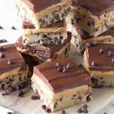 Angled view of layered Chocolate chip cookie dough, peanut butter cup filling, and chocolate ganache bars stacked on a wood cutting board.