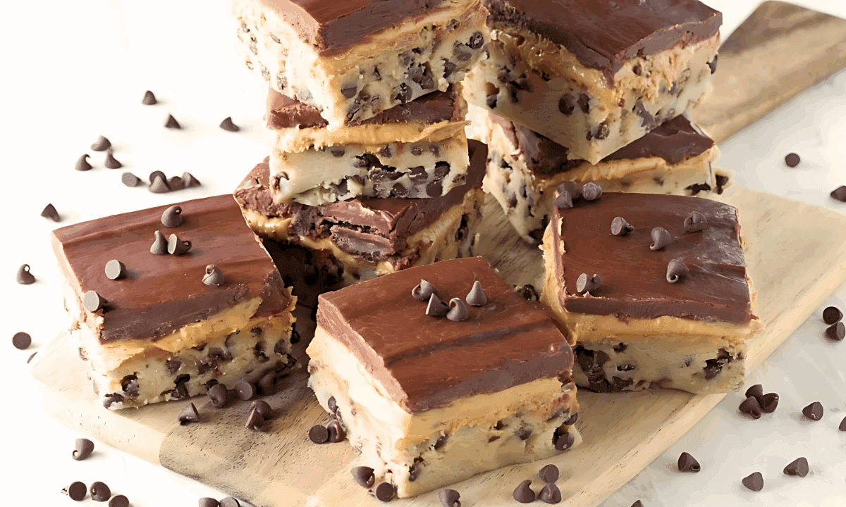 Angled view of layered Chocolate chip cookie dough, peanut butter cup filling, and chocolate ganache bars stacked on a wood cutting board.
