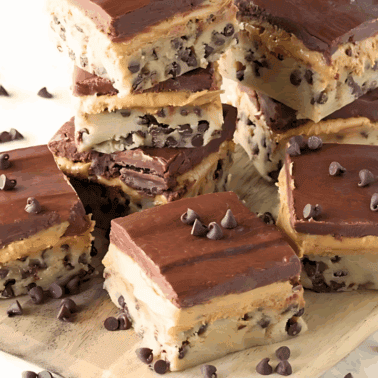 Angled view of layered Chocolate chip cookie dough, peanut butter cup filling, and chocolate ganache bars stacked on a wood cutting board.