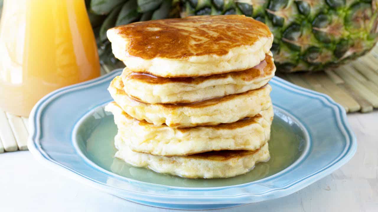 Angled view of a stack of Pineapple Pancakes on a decorative light blue plate drizzled with Coconut Syrup.