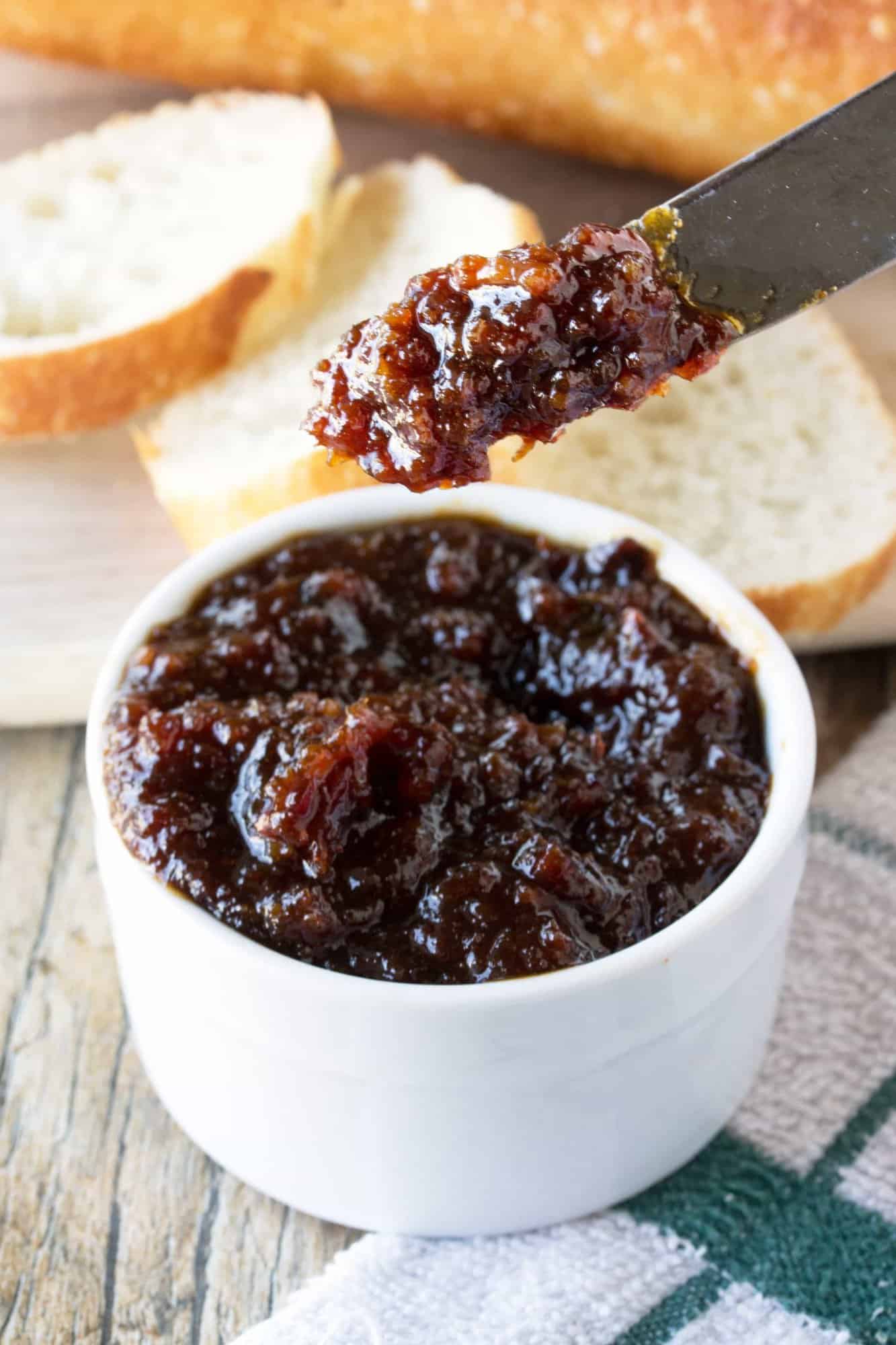 A knife with bacon jam on it above a white bowl with bacon jam in it.