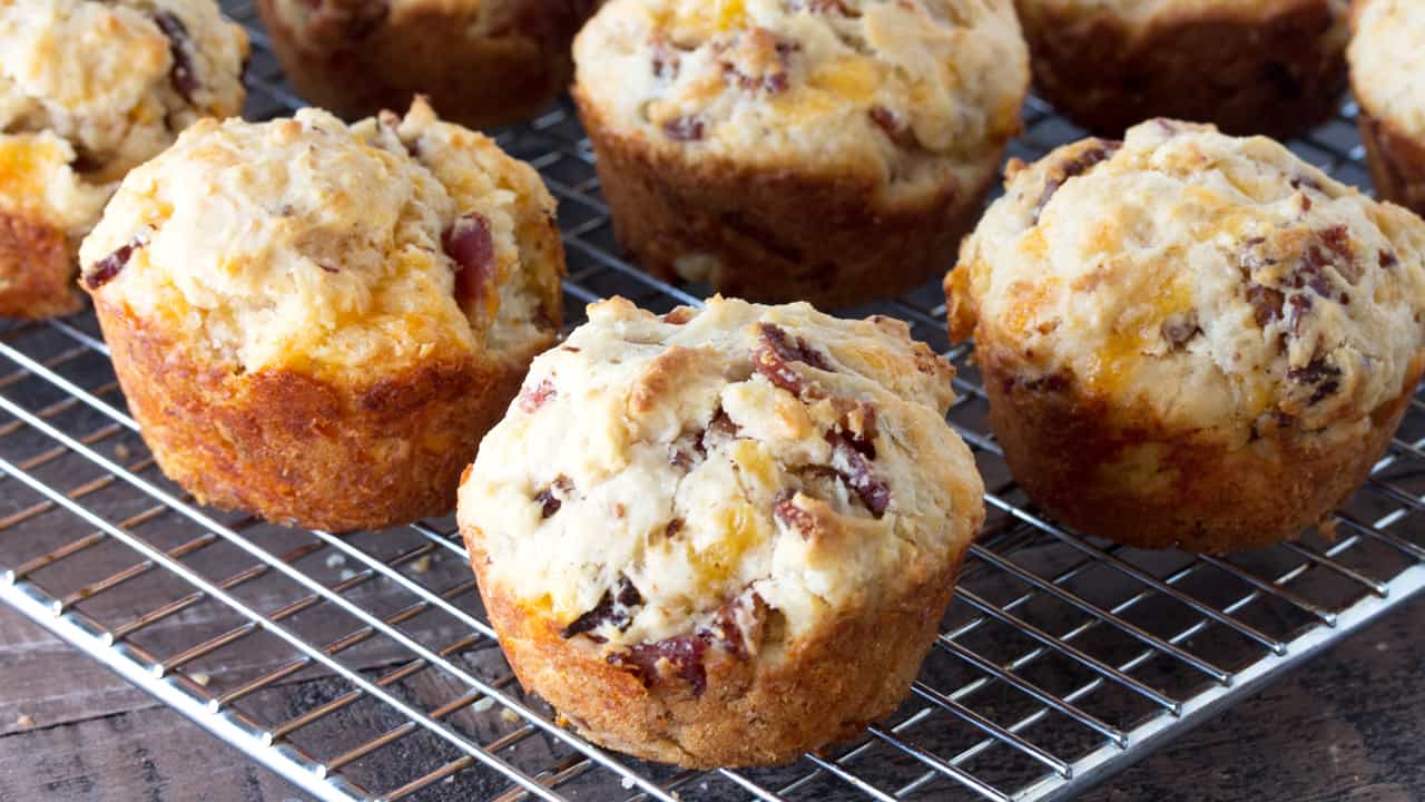 A hand is holding up a Bacon Cheddar Muffin with the rest on a cooling rack in the background.