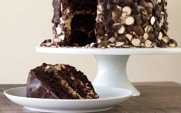 Front view of a single slice of Triple Malt Chocolate Cake on a white plate and the remainder of the cake covered with malted milk candies on a cake stand behind.
