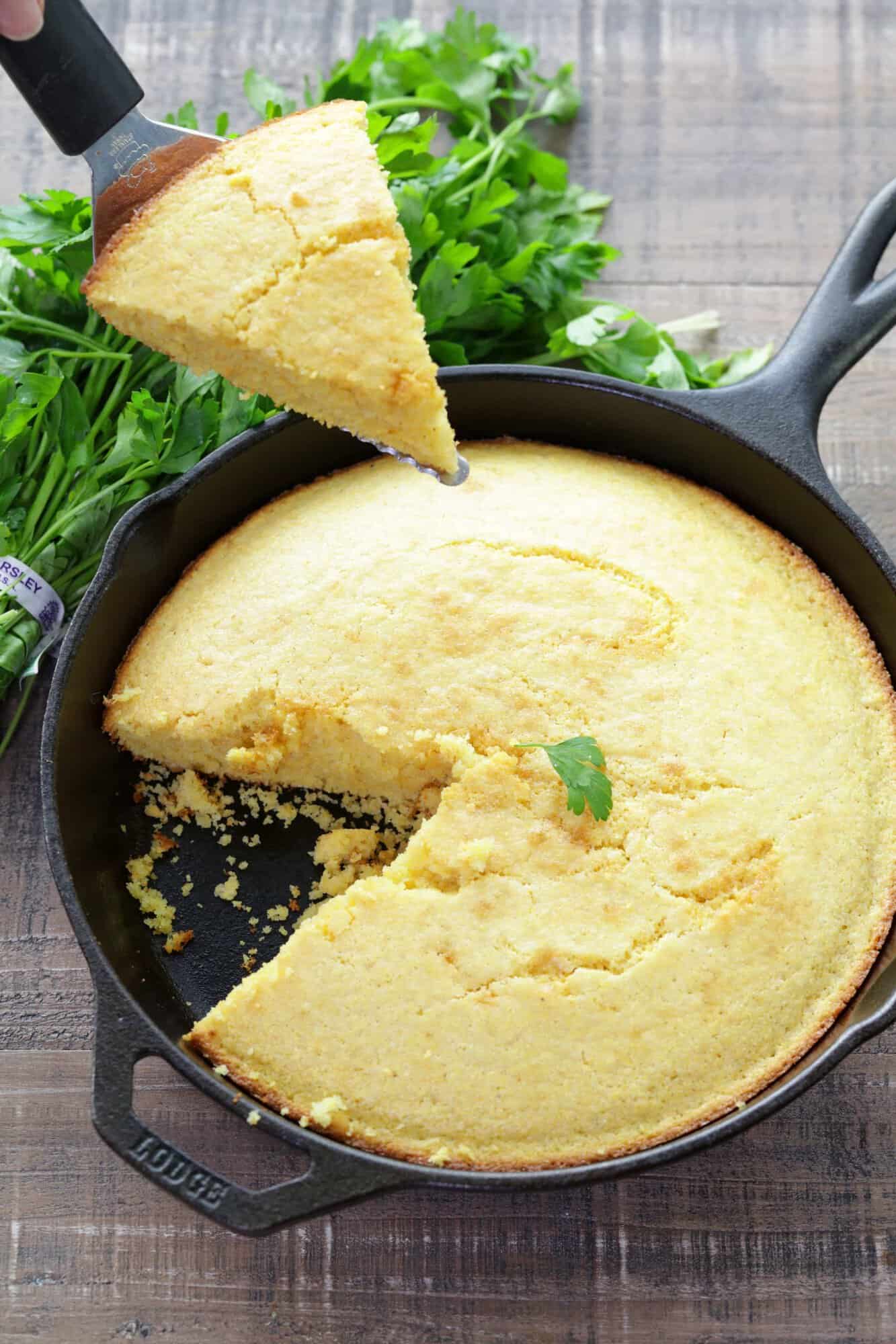 Slice of cornbread being held over skillet full of cornbread with slice removed