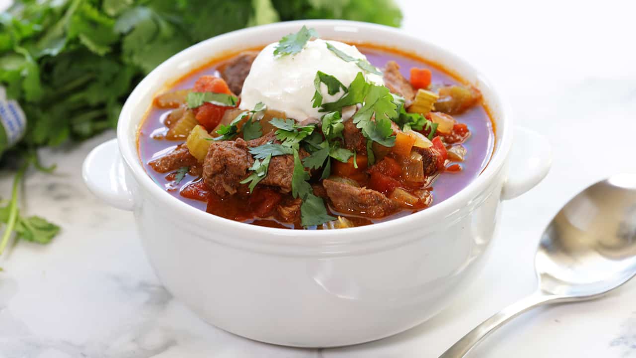 Angled view of Slow Cooker Texas Beef Chili with chunks of beef, slow cooked tomatoes, bell pepper, carrots, onion and celery and topped with sour clean, fresh cilantro.