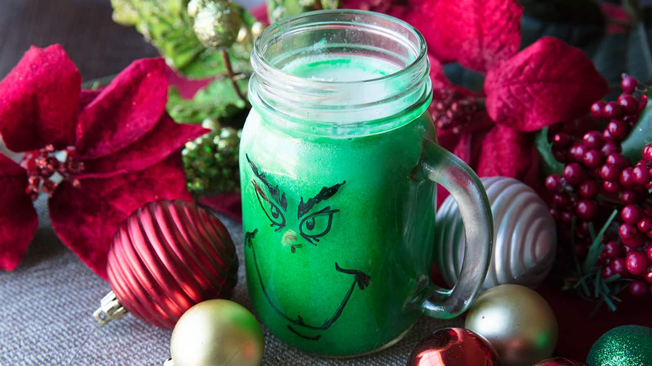 Green Grinch Punch in a mason jar glass with a handle and a grinch face drawn on it with a marker, surrounded by holiday decor.