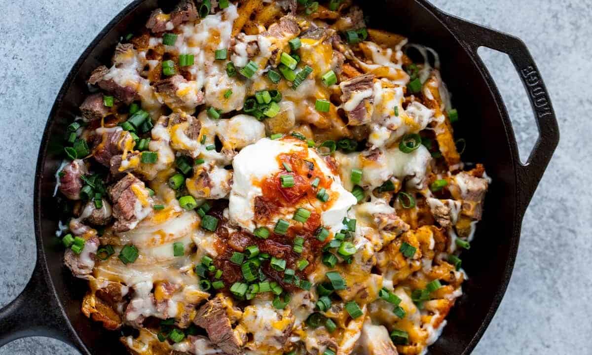Bird's eye view of Steak and Potato Nachos garnished with sour cream, green onions, and drizzled with hot sauce all in a skillet.