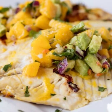 Angled view of Mango Chile Tilapia garnished with mango, avocado, red onion and parsley on a white serving dish.