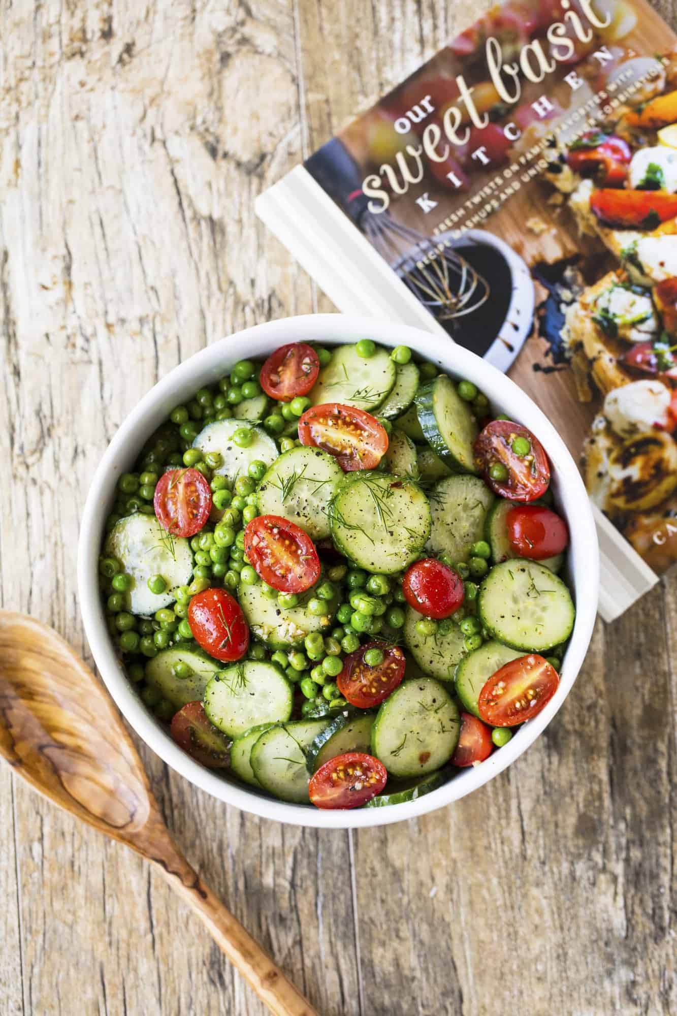 Dill Pea and Cucumber Salad in a white bowl with a wooden spoon to its left and a cookbook to its right.