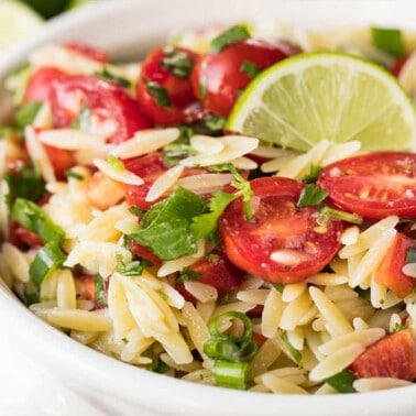 Angled view of Cilantro-Lime Orzo Pasta Salad garnished with cilantro and halved cherry tomatoes served in a white bowl.