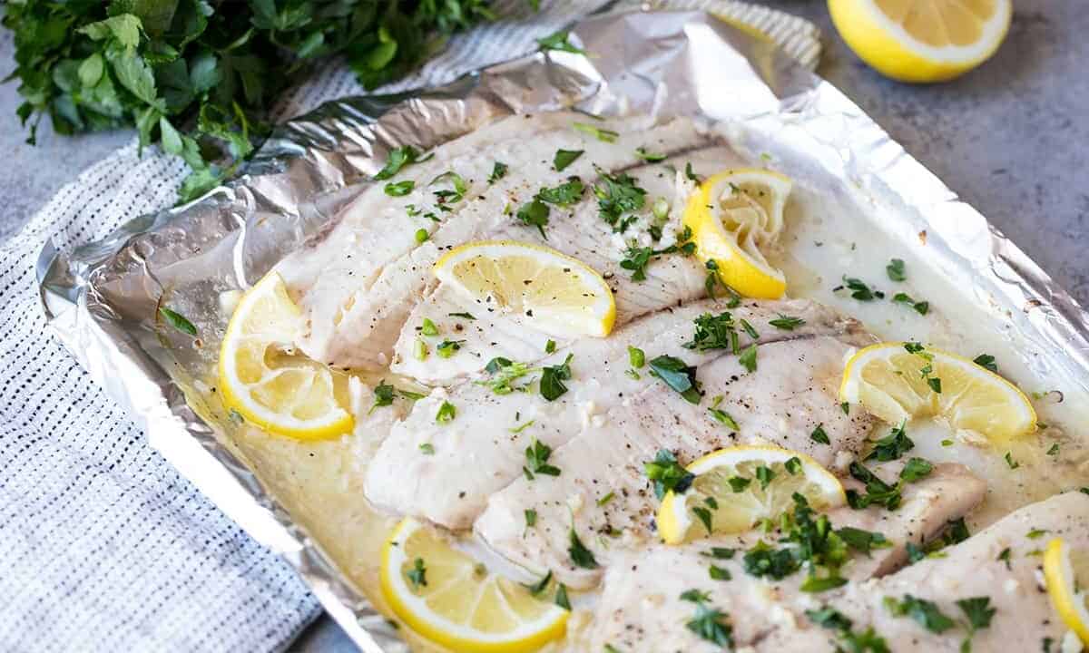 Angled view of Lemon Garlic Baked Tilapia topped with lemon slices and chopped fresh parsley on a foil lined baking sheet.