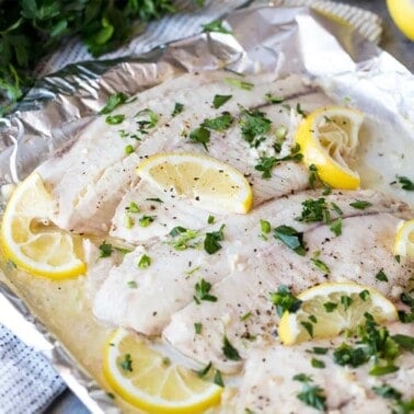 Angled view of Lemon Garlic Baked Tilapia topped with lemon slices and chopped fresh parsley on a foil lined baking sheet.