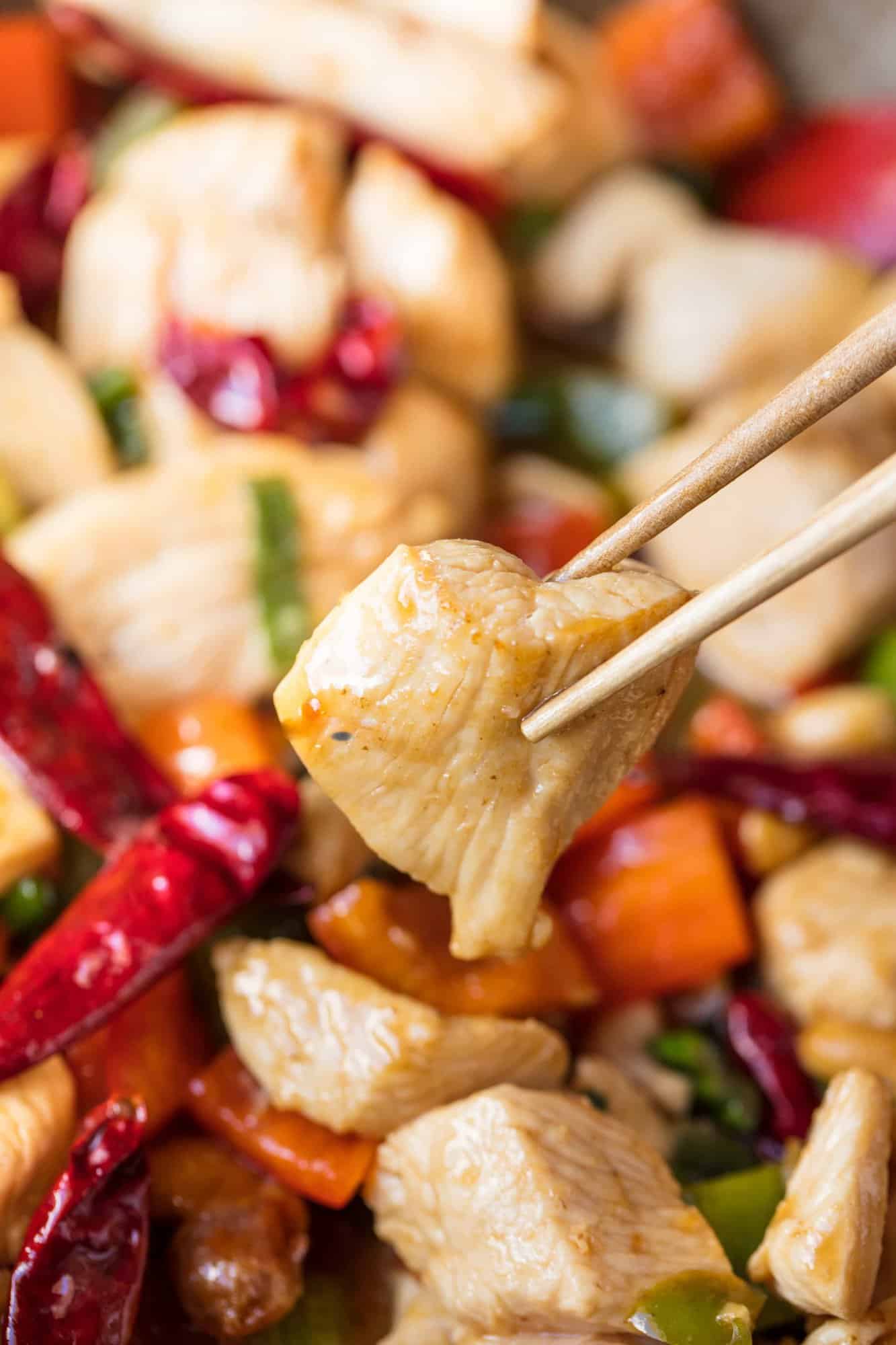 Chopsticks holding a piece of Kung Pao chicken over a bowl full of Kung Pao chicken
