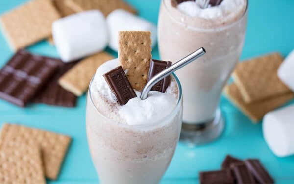 Close-up view of S'mores Frappe in a clear curved glass garnished with two pieces of Hershey's Chocolate a single graham cracker and whipped cream with a metal straw all surrounded by marshmallows, graham crackers and chocolate bars in the background.