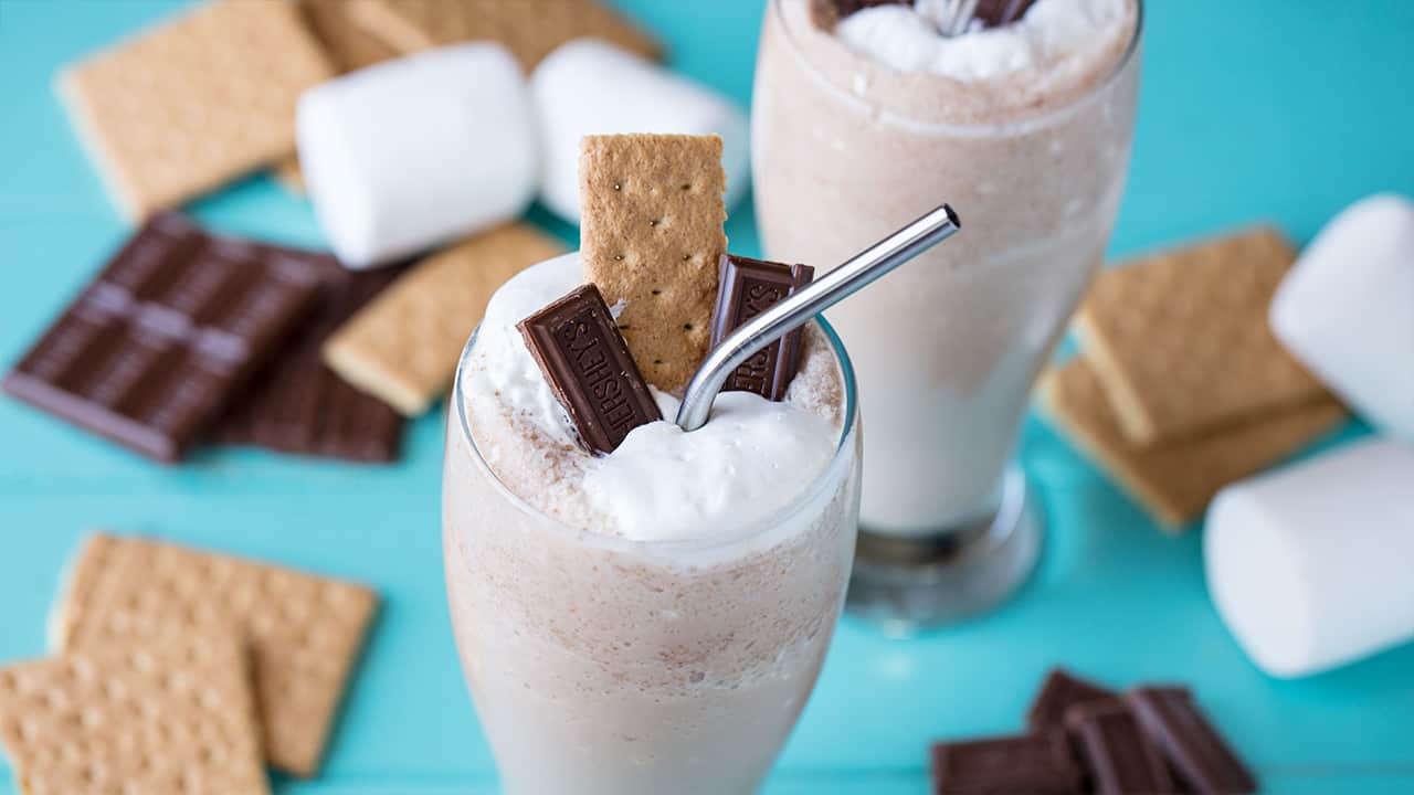 Close-up view of S'mores Frappe in a clear curved glass garnished with two pieces of Hershey's Chocolate a single graham cracker and whipped cream with a metal straw all surrounded by marshmallows, graham crackers and chocolate bars in the background.