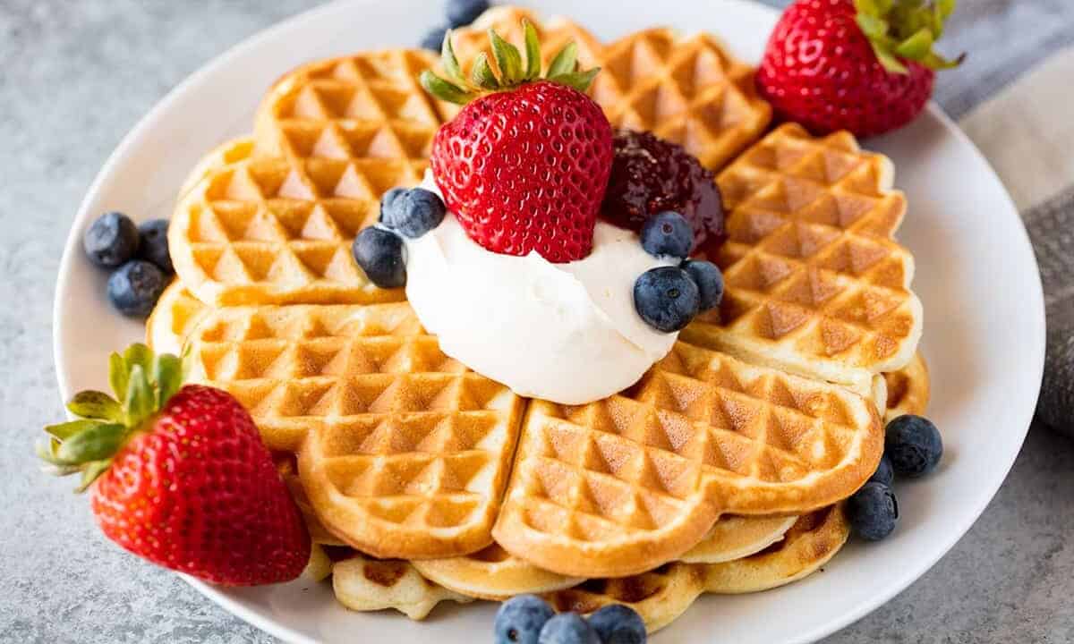 Angled view of a stack of Norwegian Waffles garnished with whipped cream, fresh strawberries, fresh blueberries, and red berry jam on a white plate all on a concrete countertop.