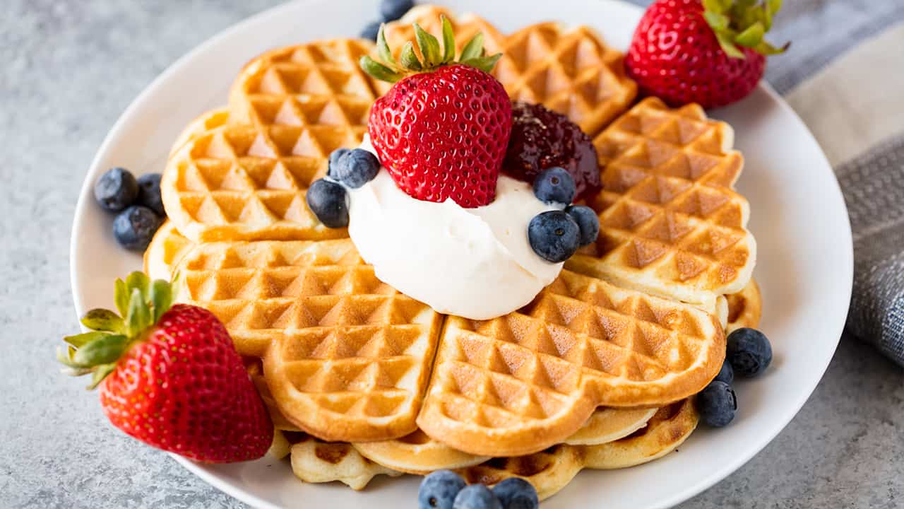 Angled view of a stack of Norwegian Waffles garnished with whipped cream, fresh strawberries, fresh blueberries, and red berry jam on a white plate all on a concrete countertop.