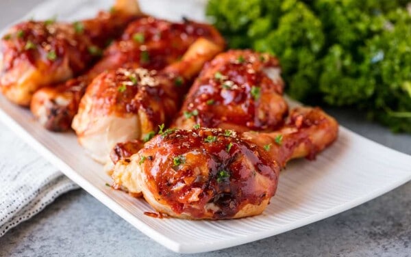 Angled view of Honey Garlic Barbecue Chicken Drumsticks garnished with parsley, lined up on a white serving platter.