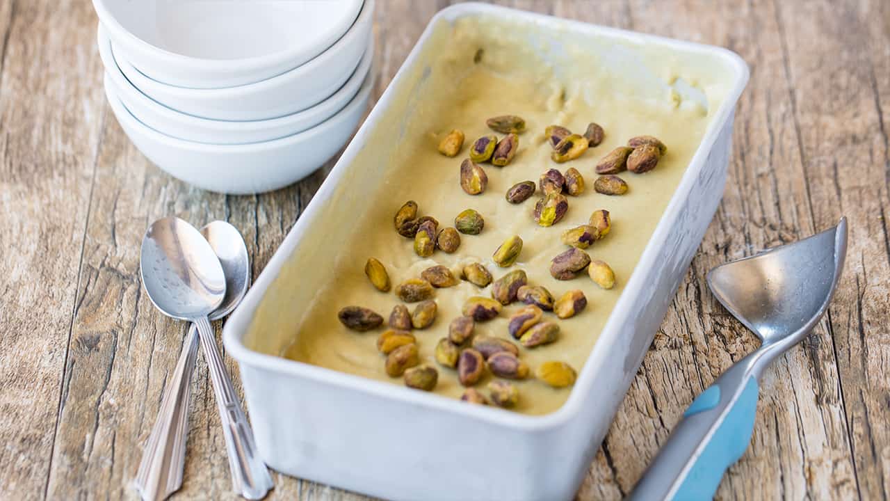Angled view of Pistachio Gelato in stainless steel bread pan garnished with whole pistachios with stack of white serving bowls on worn wood table top.
