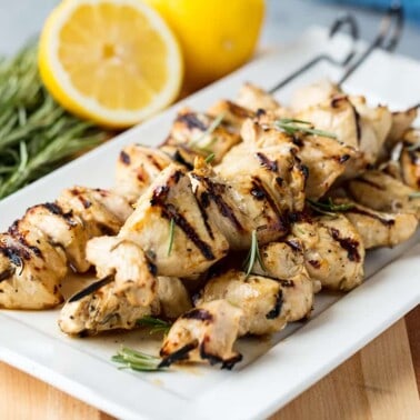 Angled view of Rosemary Ranch Chicken Kabobs stacked on a white serving platter garnished with rosemary leaves and surrounded by sprigs of rosemary and lemon halves all on a wood cutting board.
