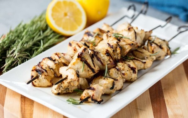 Angled view of Rosemary Ranch Chicken Kabobs stacked on a white serving platter garnished with rosemary leaves and surrounded by sprigs of rosemary and lemon halves all on a wood cutting board.