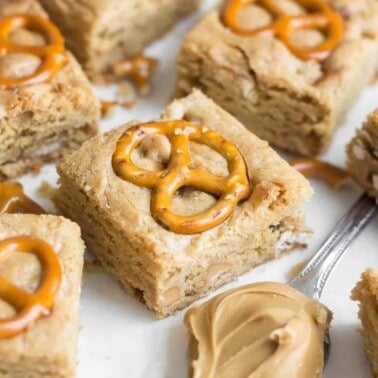 Peanut butter pretzel blondie squares each topped with a pretzel and a spoonful of peanut butter sitting on parchment paper