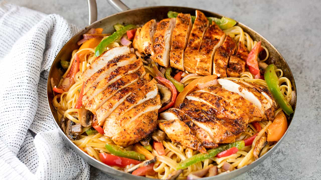 Angled view of Cajun spiced chicken breasts sliced and served on a bed of linguini with grilled bell peppers, mushrooms and red onion in a skillet
