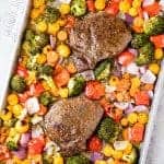 Two steaks are surrounded by roasted broccoli, cherry tomatoes, bell peppers and red onion on a sheet pan
