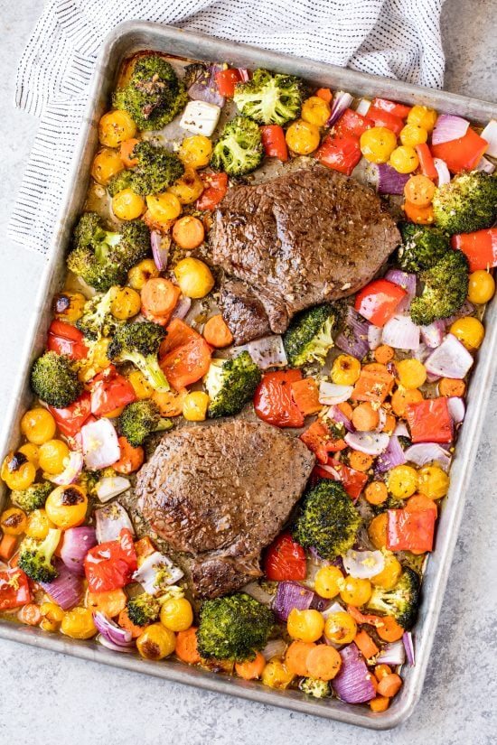 Two steaks are surrounded by roasted broccoli, cherry tomatoes, bell peppers and red onion on a sheet pan