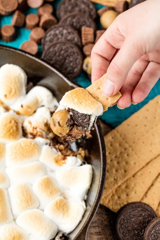 Rachel scoops a bite of million dollar skillet s'mores with a graham cracker from a cast iron skillet full of marshmallow, cookie dough, Oreo, and other melty goodness