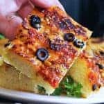 A slice of easy zesty focaccia topped with marinara sauce, sliced olives, Italian seasoning, parmesan cheese and pine nuts
