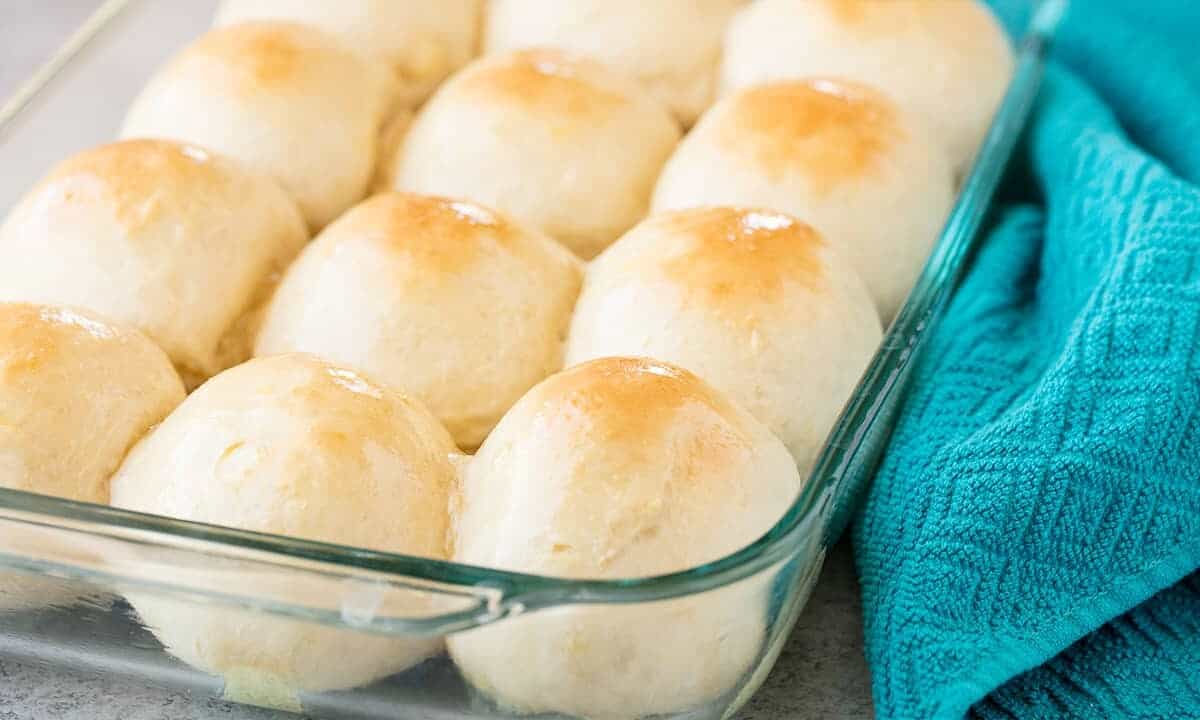 Angled view of Hawaiian Sweet Rolls glistening with warm butter nestled in glass casserole dish with a teal colored tea towel to the side.