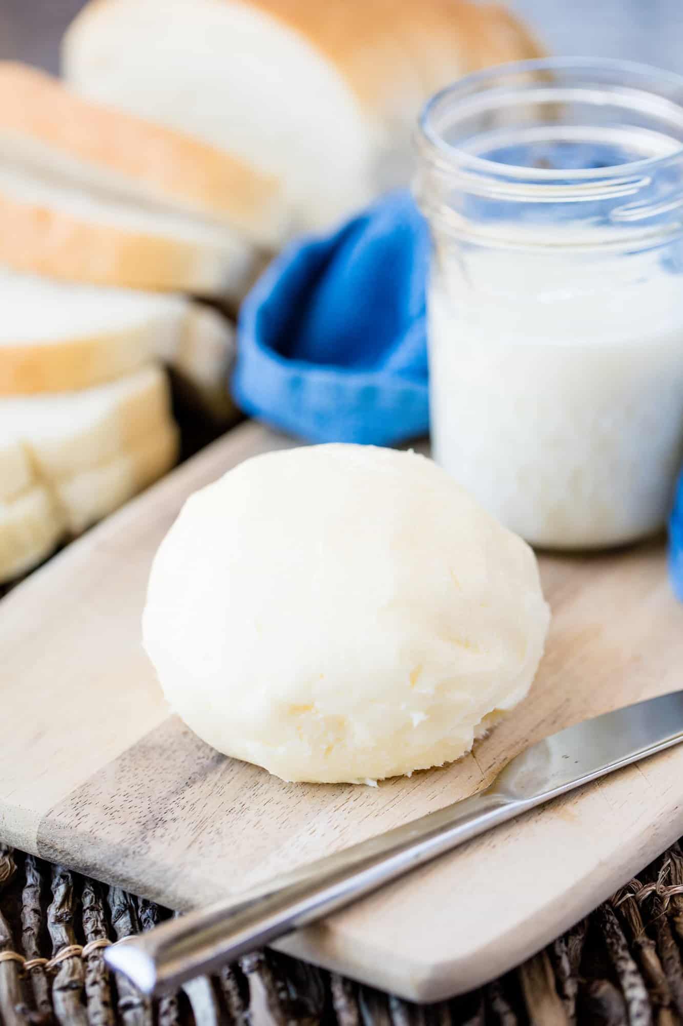 A ball of homemade butter on a cutting board with a butter knife and a jar of cream in the background
