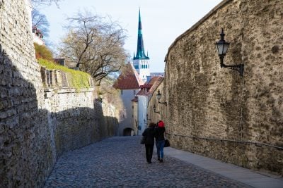 Two people stroll down a cobblestone street surrounded by rock walls and plants dripping over the side. A church steeple in the distance