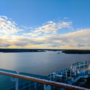 A sweeping view across the cruise ship with ocean, the glimpse of land (Nynashamn) and clouds lit by the sun