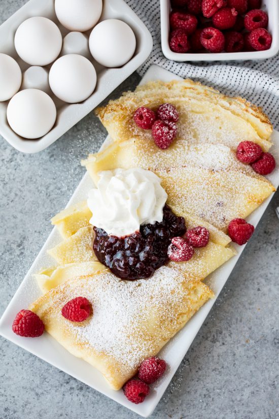 Fluffy Swedish Pancakes served on a platter with fresh raspberries, jam, a dollop of whipped cream and a dusting of powdered sugar