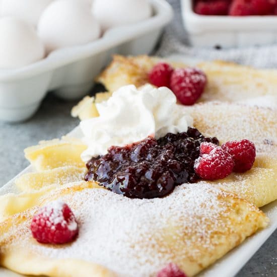 Fluffy Swedish Pancakes served on a platter with fresh raspberries, jam, a dollop of whipped cream and a dusting of powdered sugar
