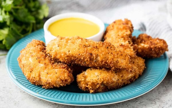 Angled view of a stack of Cajun Fried Chicken Strips with a ramekin of honey mustard sauce all on a teal dinner plate.