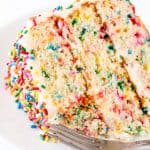 A close up of a slice of Funfetti Birthday Cake with a bite taken out of it on a white plate with a fork on it.