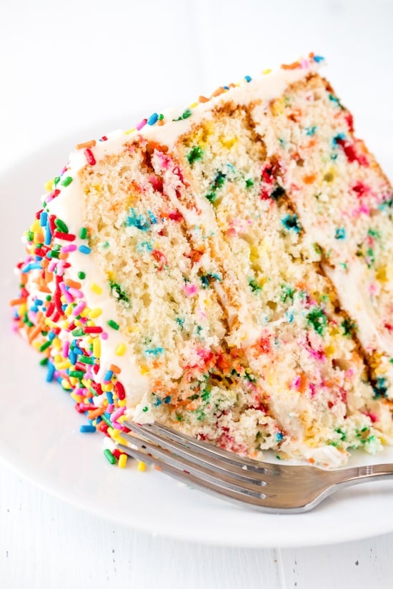 A close up of a slice of Funfetti Birthday Cake with a bite taken out of it on a white plate with a fork on it.