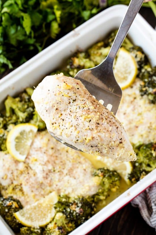 Lemon chicken being held by a spatula over a pan full of lemon garlic chicken broccoli bake. 