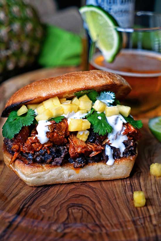 Pineapple Chile Pulled Pork Sandwiches sitting on a wood counter top with a drink in the background.