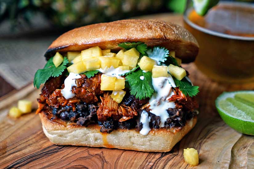 This slow simmered Pineapple Guajillo Chile Pulled Pork is stuffed in a ciabatta roll, layered on top of mashed black beans, a drizzle of Mexican crema, cilantro and diced pineapple. keviniscooking.com