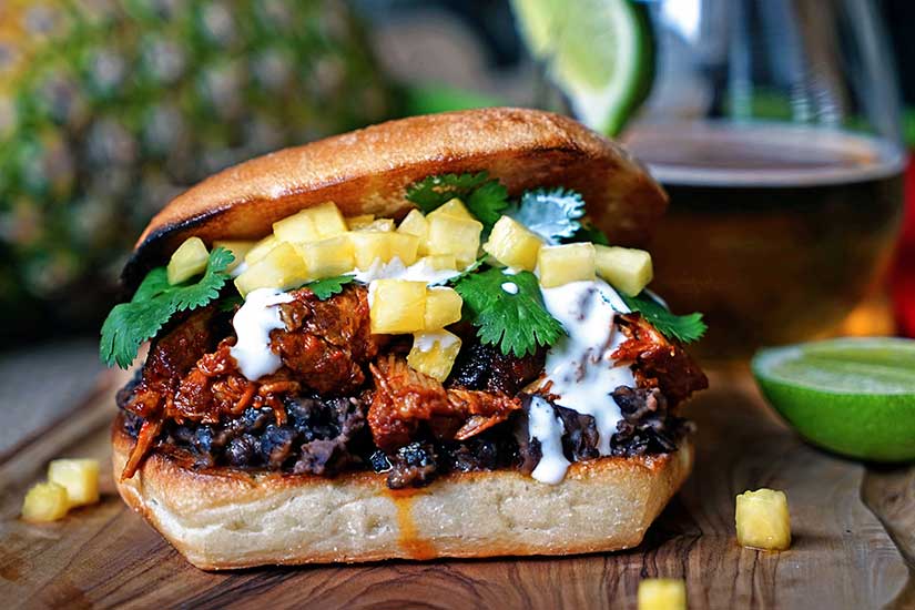 Close up of a Pineapple Chile Pulled Pork Sandwiches sitting on a wood counter top with a drink in the background.