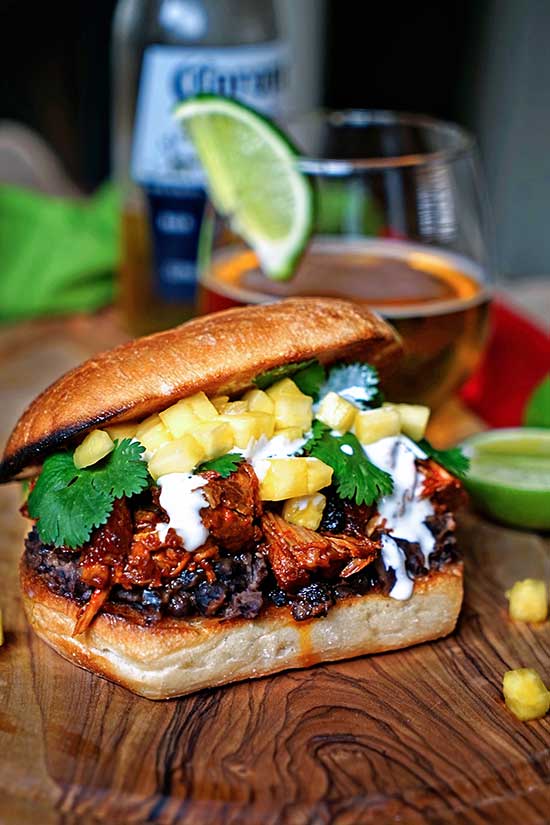 Angeled shot of a Pineapple Chile Pulled Pork Sandwiches sitting on a wood countertop with a drink in the background.