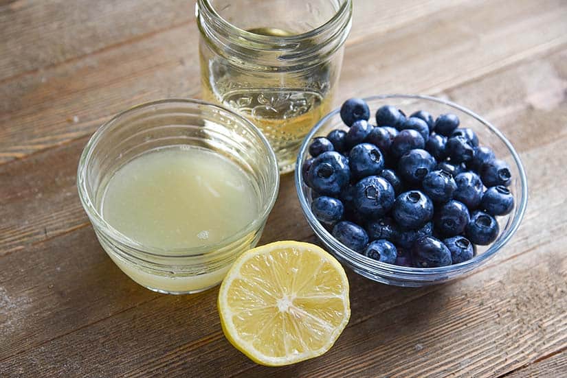 Glass of syrup water, bowl of lemon juice, bowl of blueberries, and a half a lemon on a wood countertop.