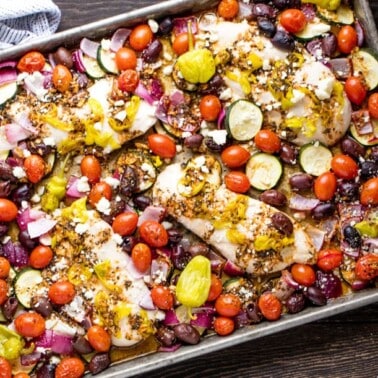 Sheet Pan Greek Chicken and Veggie Bake with cherry tomatoes, olives, red onion, zucchini, pepperoncini and topped with crumbled feta cheese on a sheet pan